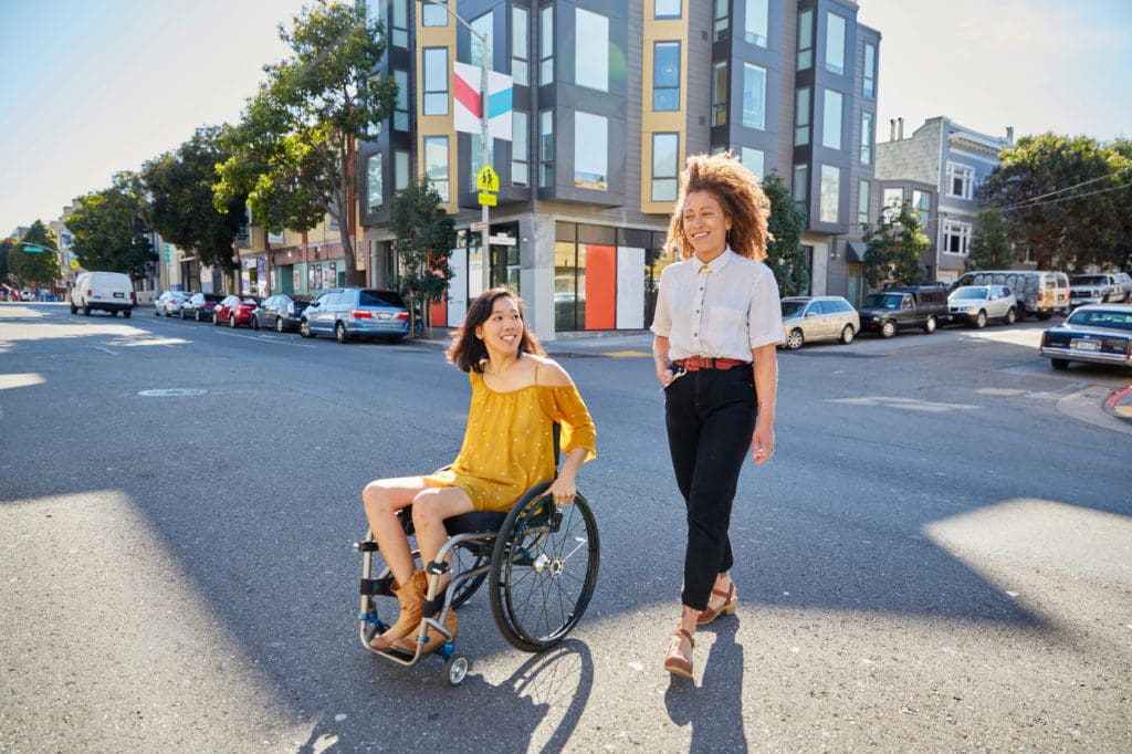 Two younger adults rolling and walking in the middle of an urban setting with a modern high rise building in the background. One person is using a wheelchair.