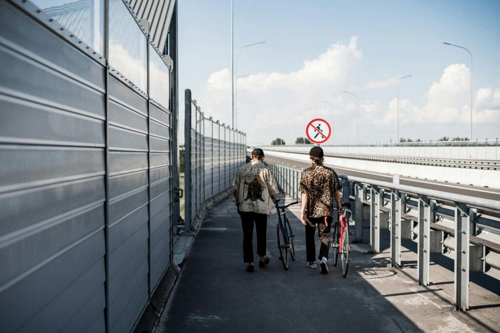 Two youth with bicycles in hand walk down a pedestrian path next to a highway. There is a no walking symbol in the horizon.