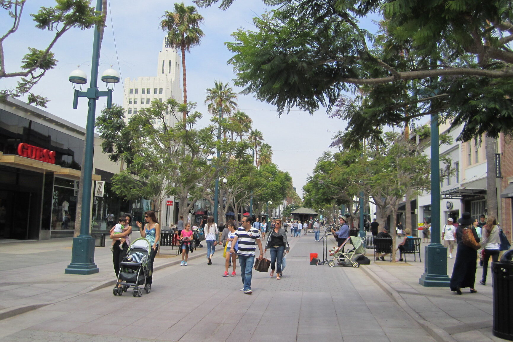 Pedestrian street with shops, trees, and people walking, shopping, and relaxing. Stores and a tall art deco building are visible in the background.