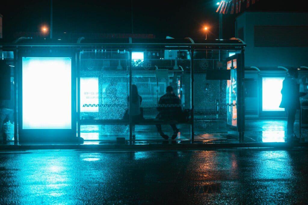 Una parada de autobús con luz azul oscura y dos personas sentadas en un banco, esperando.