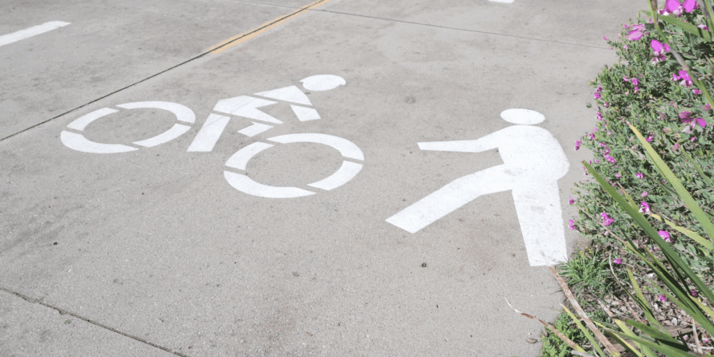 White stenciled pedestrian and bicyclist images on a gray sidewalk with a floral bush on the right side