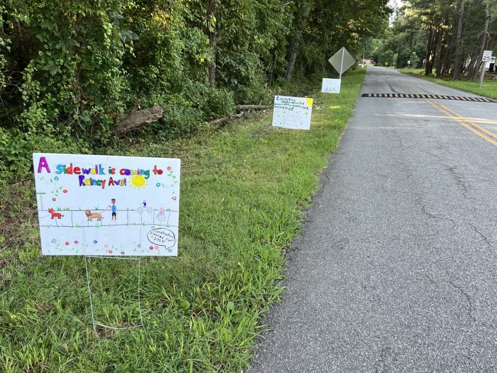 signs depicting and calling for sidewalks on the side of a road with a speed bump